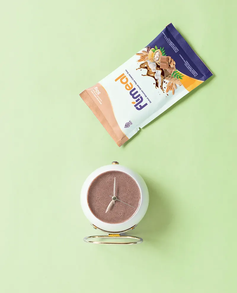 Sachet Flimeal dan minuman Flimeal cokelat di dalam jam meja, latar hijau